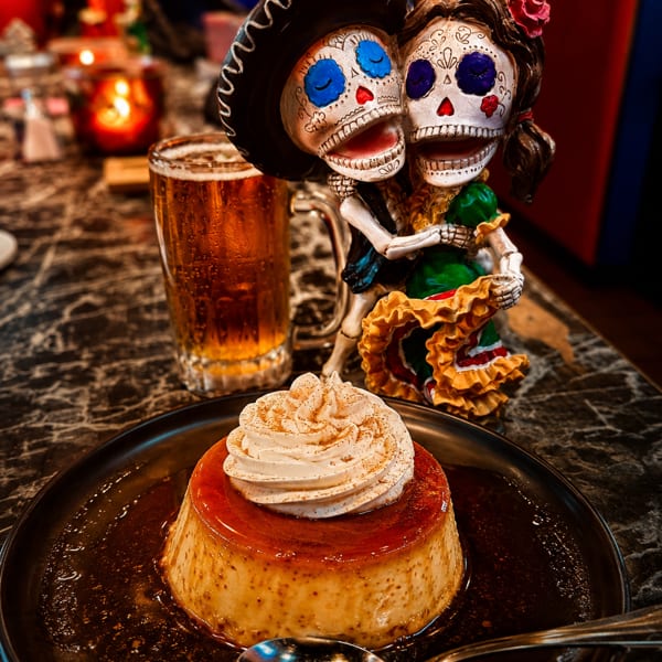 Flan with whipped cream and cinnamon, a beer, and Day of the Dead figurines on a dark table.