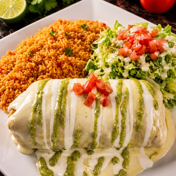 A large burrito covered in white and green sauce, served with rice and a side salad.