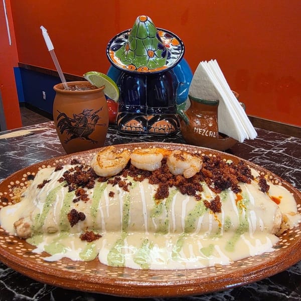 A large burrito covered in white and green sauce, topped with shrimp and chorizo, on a brown plate.